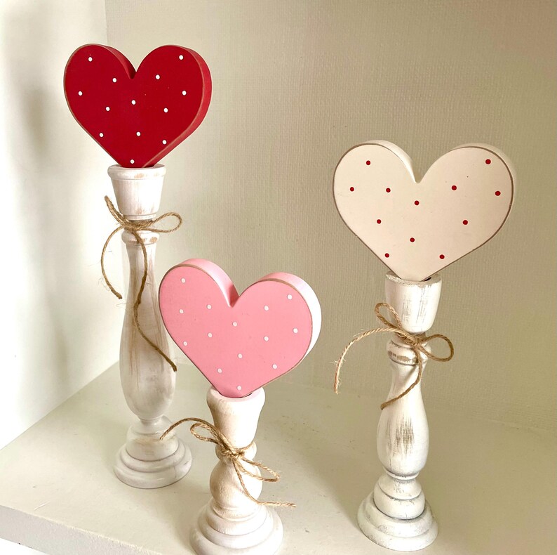 May include: Three wooden heart decorations on white candlestick holders. The hearts are red, pink, and white, each with a polka dot pattern. The candlesticks are tied with twine, creating a rustic, Valentine's Day theme.