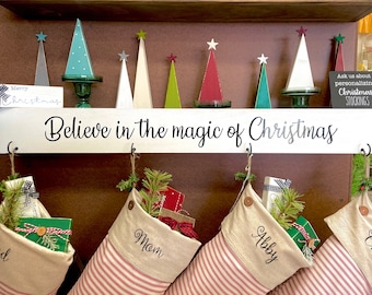 May include: A white wooden sign with the text "Believe in the magic of Christmas" hangs above four personalized Christmas stockings with red and white stripes. The stockings are labeled "Dad", "Mom", "Abby", and "Charl".
