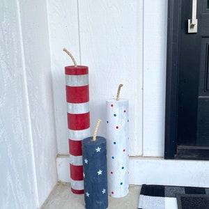 May include: Three wooden cylinders painted to look like fireworks. The largest cylinder is red and white stripes, the middle cylinder is blue with white stars, and the smallest cylinder is white with red and blue polka dots. Each cylinder has a brown twine attached to the top.
