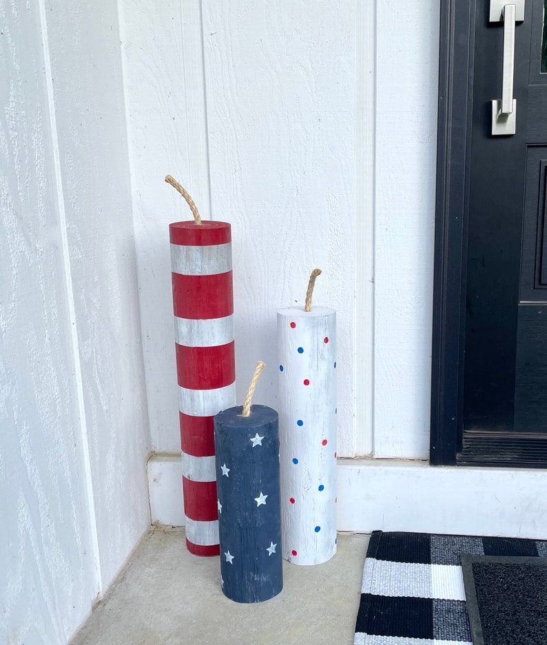 May include: Three wooden cylinders painted to look like fireworks. The largest cylinder is red and white stripes, the middle cylinder is blue with white stars, and the smallest cylinder is white with red and blue polka dots. Each cylinder has a brown twine attached to the top.