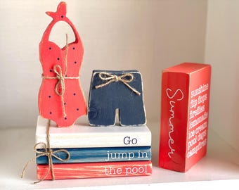 May include: Wooden summer-themed decor featuring a red swimsuit with black polka dots, navy blue shorts, and a red box with "Summer" and summer-related words. The swimsuit and shorts are tied with twine. The words "Go jump in the pool" are on the stacked books.