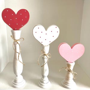 May include: Three wooden heart-shaped decorations on white candlestick holders. The hearts are red, white, and pink, each with white or red polka dots. The candlesticks are tied with twine bows, creating a charming, romantic display.