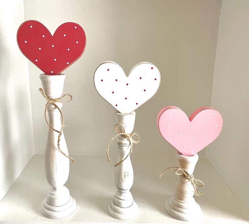 May include: Three wooden heart-shaped decorations on white candlestick holders. The hearts are red, white, and pink, each with white or red polka dots. The candlesticks are tied with twine bows, creating a charming, romantic display.