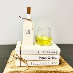 May include: A stack of three white books with the words "Chardonnay", "Pinot Grigio", and "Sauvignon Blanc" printed on them. A wooden wine bottle with the word "Wine" printed on it sits on top of the books. A glass of white wine sits next to the bottle.