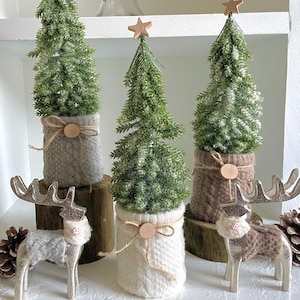 May include: Three miniature artificial Christmas trees with wooden star toppers, set in knitted fabric pots. Two silver reindeer figurines and pinecones complete the holiday decor. The trees are on wooden bases.