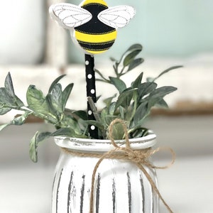 May include: A white ceramic planter with a distressed finish and vertical lines. The planter has a brown twine bow and a black and white striped stick with a wooden bee on top. The bee is yellow and black with white wings.