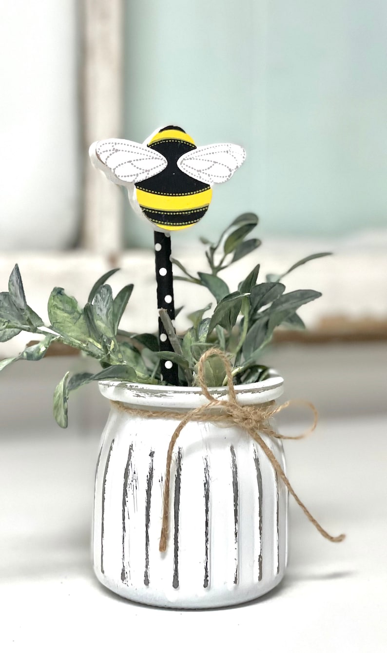May include: A white ceramic planter with a distressed finish and vertical lines. The planter has a brown twine bow and a black and white striped stick with a wooden bee on top. The bee is yellow and black with white wings.