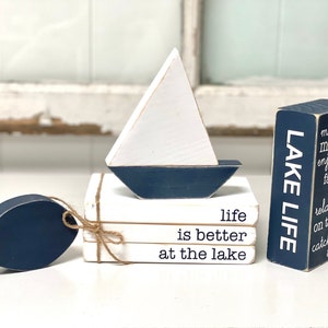 May include: A set of three white wooden blocks stacked on top of each other with the words "life is better at the lake" written in black. A small blue and white wooden sailboat sits on top of the blocks. A blue wooden fish is on the left side of the image. A wooden block with the words "LAKE LIFE" written in white on a blue background is on the right side of the image.