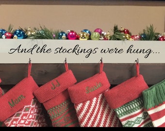 May include: A white wooden box with the text "And the stockings were hung..." on the front. The box has hooks on the bottom for hanging stockings. Five stockings are hanging from the hooks. The stockings are red and green with white stripes and have names embroidered on them: Mom, JJ, Susie, and two others.