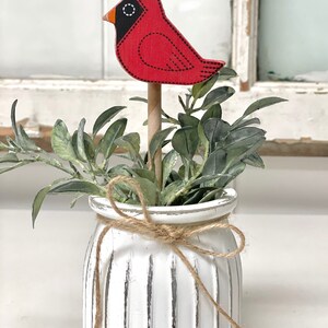 May include: A white ceramic planter with a rustic twine bow, filled with green foliage and a red cardinal bird figurine.