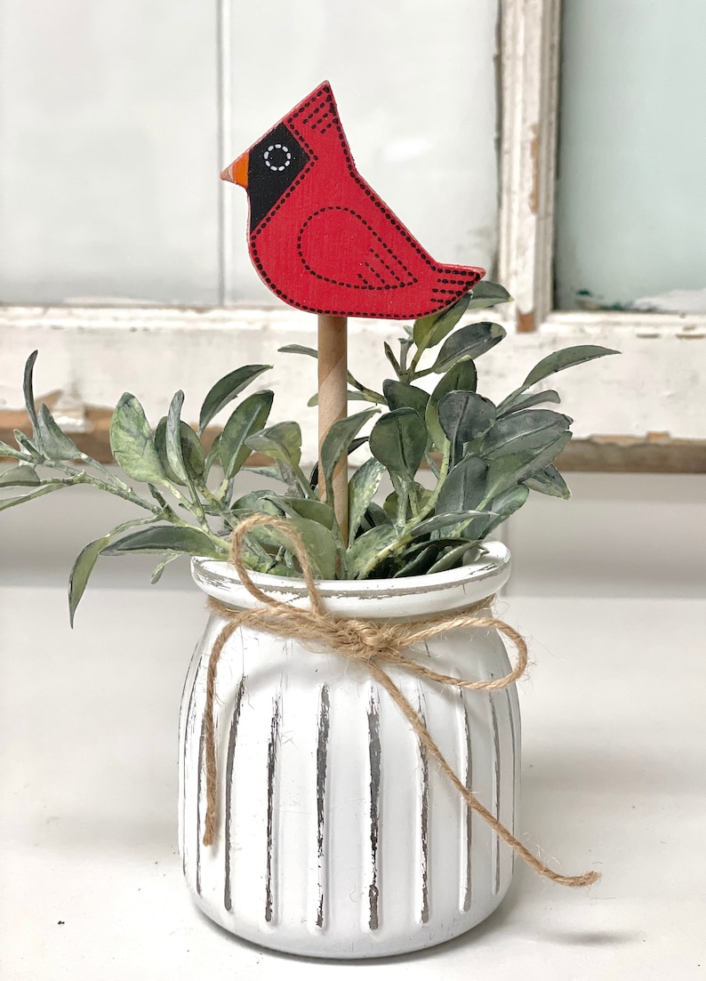 May include: A white ceramic planter with a rustic twine bow, filled with green foliage and a red cardinal bird figurine.