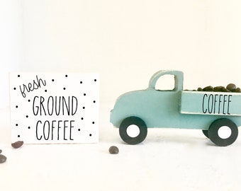 May include: A white wooden sign with black polka dots and the text "fresh ground coffee" in black script. A light blue toy pickup truck with a white bed filled with brown coffee beans. The bed of the truck has the word "coffee" written in black.