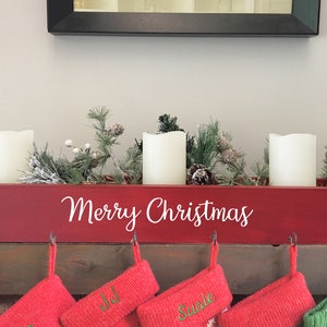 May include: A red wooden Christmas centerpiece with the words "Merry Christmas" in white script. It holds three white pillar candles, pine branches, and pinecones. Below are red Christmas stockings.