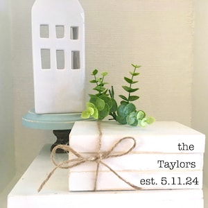 May include: A stack of three white books with the text "the Taylors est. 5.11.24" printed on the top book. The books are tied together with twine and decorated with a small sprig of greenery.
