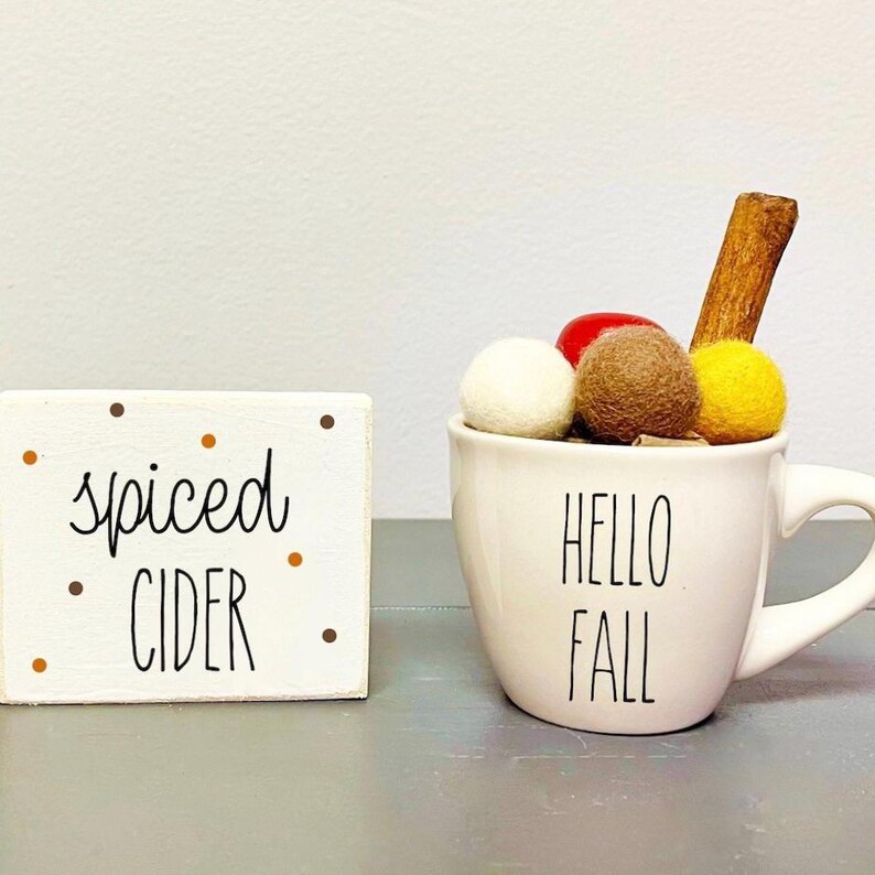 Pode incluir: Caneca de cer&acirc;mica branca com o texto "Hello Fall" nela. A caneca est&aacute; cheia de bolas de feltro brancas, marrons e amarelas e um pau de canela. Um pequeno letreiro de madeira com o texto "spiced cider" est&aacute; no fundo.
