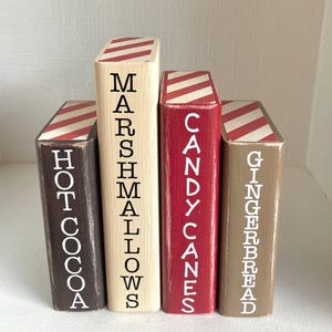 K&ouml;nnte beinhalten: Vier Holzbl&ouml;cke, die in verschiedenen Farben bemalt sind und jeweils einem Buch &auml;hneln. Die Bl&ouml;cke sind mit den W&ouml;rtern "Hot Cocoa", "Marshmallows", "Candy Canes" und "Gingerbread" in wei&szlig;en Buchstaben beschriftet. Die Oberseiten der Bl&ouml;cke haben ein rot-wei&szlig; gestreiftes Design.