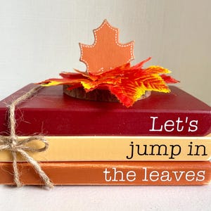 May include: A stack of three wooden blocks painted in fall colors: burgundy, yellow, and orange. The blocks are tied with twine and topped with a wooden leaf and a decorative fall leaf. The text on the blocks reads "Let's jump in the leaves."