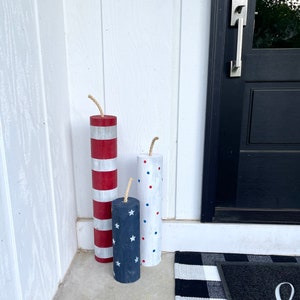 May include: Three wooden firecracker decorations painted in red, white, and blue with stars and polka dots. The firecrackers are standing on a concrete porch in front of a black door.