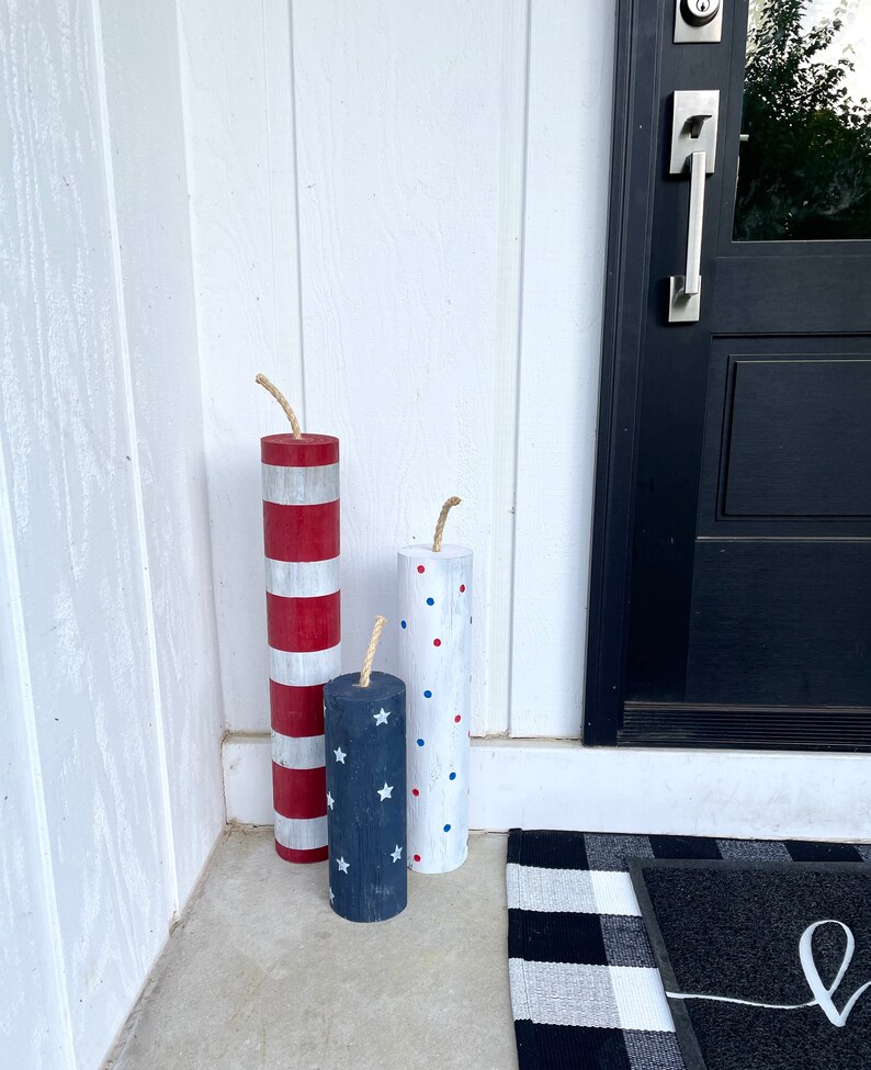 May include: Three wooden firecracker decorations painted in red, white, and blue with stars and polka dots. The firecrackers are standing on a concrete porch in front of a black door.