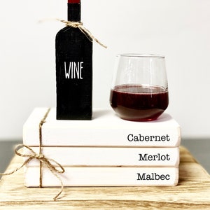 May include: A decorative black wine bottle with the word "WINE" in white, a stemless wine glass filled with red wine, and three white books labeled "Cabernet", "Merlot", and "Malbec". The bottle is tied with twine.