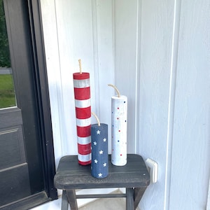 May include: Three wooden decorative firecrackers painted in red, white, and blue patriotic colors. The firecrackers are decorated with stars and polka dots. They are sitting on a black wooden stool.