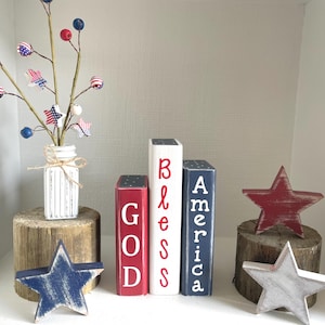 May include: A patriotic display featuring wooden blocks that spell out "GOD BLESS AMERICA." Accompanying the blocks are wooden stars in red, white, and blue, and a vase with decorative branches. The arrangement is set on wooden bases.
