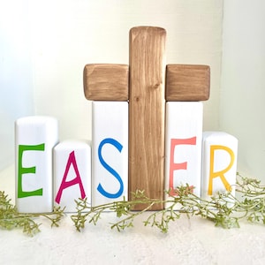 May include: Wooden blocks spelling out the word "EASTER" in colorful letters. The blocks are arranged in a row with a wooden cross behind them. The cross is made of two pieces of wood, one vertical and one horizontal. The blocks are painted white and the letters are painted in different colors. The blocks are sitting on a white surface.