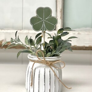 May include: A white ceramic planter with a rustic finish and vertical lines. The planter is tied with twine and holds a green plant with a green shamrock cutout.