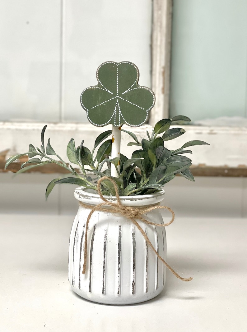 May include: A white ceramic planter with a rustic finish and vertical lines. The planter is tied with twine and holds a green plant with a green shamrock cutout.