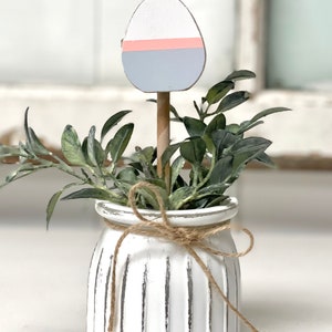 May include: A white ceramic planter with a rustic finish and a brown twine bow. The planter is filled with green foliage and a wooden egg painted in shades of white, pink, and blue.
