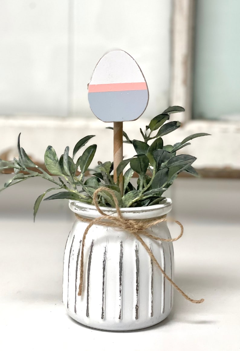 May include: A white ceramic planter with a rustic finish and a brown twine bow. The planter is filled with green foliage and a wooden egg painted in shades of white, pink, and blue.