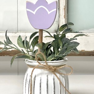 May include: A white ceramic planter with a rustic finish and a brown twine bow. The planter is filled with green foliage and a purple wooden tulip.