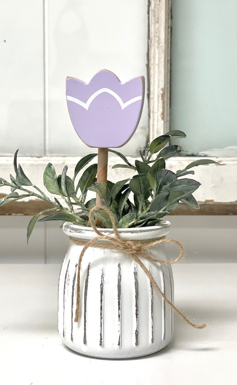 May include: A white ceramic planter with a rustic finish and a brown twine bow. The planter is filled with green foliage and a purple wooden tulip.