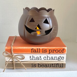 May include: A brown pumpkin-shaped candle holder with a carved face sits atop three decorative wooden books. The books are tied with twine and painted orange, white, and brown, with the phrases "fall is proof that change is beautiful."