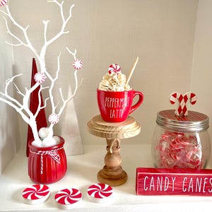 May include: A festive Christmas display featuring a red mug with "Peppermint Latte" text, a jar of candy canes, and a white tree adorned with peppermint candies. A wooden sign reads "CANDY CANES".