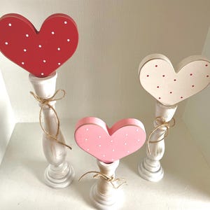 May include: Three wooden heart decorations in red, pink, and cream, each with white or red polka dots. The hearts are mounted on white candlestick-style bases and tied with twine. A charming, decorative accent.