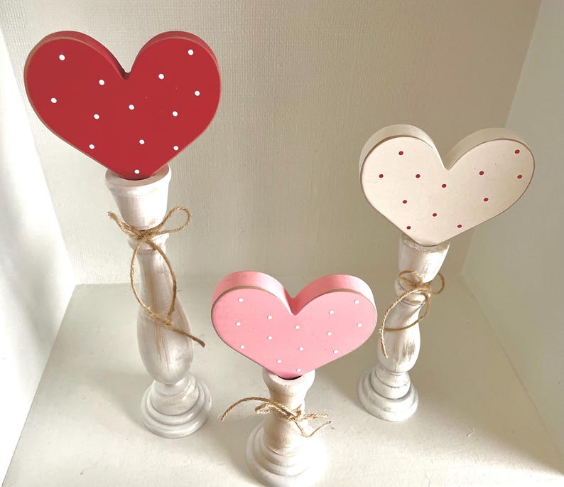 May include: Three wooden heart decorations in red, pink, and cream, each with white or red polka dots. The hearts are mounted on white candlestick-style bases and tied with twine. A charming, decorative accent.