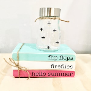 May include: Three stacked books with the words "hello summer", "fireflies", and "flip flops" printed on them. A clear glass jar with a silver lid and firefly silhouettes is on top of the books.