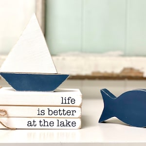 May include: A set of three white wooden blocks stacked on top of each other with the text "life is better at the lake" written in blue. A blue wooden sailboat and a blue wooden fish are on the table next to the blocks.