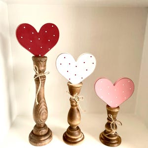 Pode incluir: Tr&ecirc;s decora&ccedil;&otilde;es de madeira em forma de cora&ccedil;&atilde;o. Cada cora&ccedil;&atilde;o &eacute; montado em um casti&ccedil;al de madeira. Os cora&ccedil;&otilde;es s&atilde;o vermelhos, brancos e rosa, cada um com bolinhas brancas. Os casti&ccedil;ais marrons s&atilde;o amarrados com barbante.