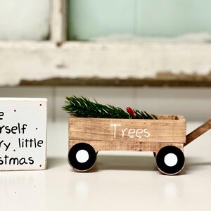 May include: A white wooden sign with the text "Have yourself a merry little Christmas."  A miniature wooden wagon with a green pine branch inside and the word "Trees" written on the side.