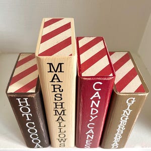 K&ouml;nnte beinhalten: Vier dekorative Holzbl&ouml;cke mit rot-wei&szlig; gestreiften Oberseiten. Die Bl&ouml;cke sind mit "Hot Cocoa", "Marshmallows", "Candy Canes" und "Gingerbread" in wei&szlig;er Schrift beschriftet. Die Bl&ouml;cke sind in verschiedenen Braun- und Rott&ouml;nen gehalten.
