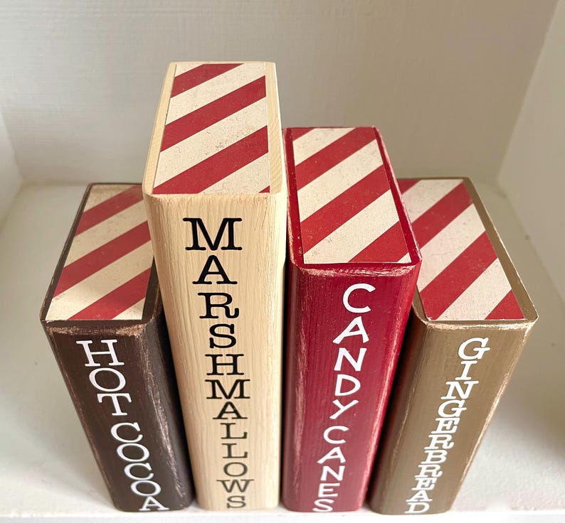 K&ouml;nnte beinhalten: Vier dekorative Holzbl&ouml;cke mit rot-wei&szlig; gestreiften Oberseiten. Die Bl&ouml;cke sind mit "Hot Cocoa", "Marshmallows", "Candy Canes" und "Gingerbread" in wei&szlig;er Schrift beschriftet. Die Bl&ouml;cke sind in verschiedenen Braun- und Rott&ouml;nen gehalten.