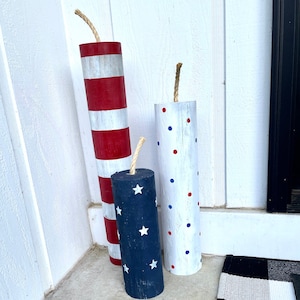 May include: Three wooden decorative firecrackers with red, white, and blue paint. The firecrackers are decorated with stripes, polka dots, and stars.