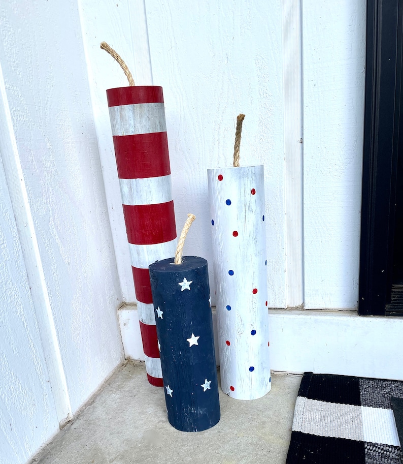May include: Three wooden decorative firecrackers with red, white, and blue paint. The firecrackers are decorated with stripes, polka dots, and stars.