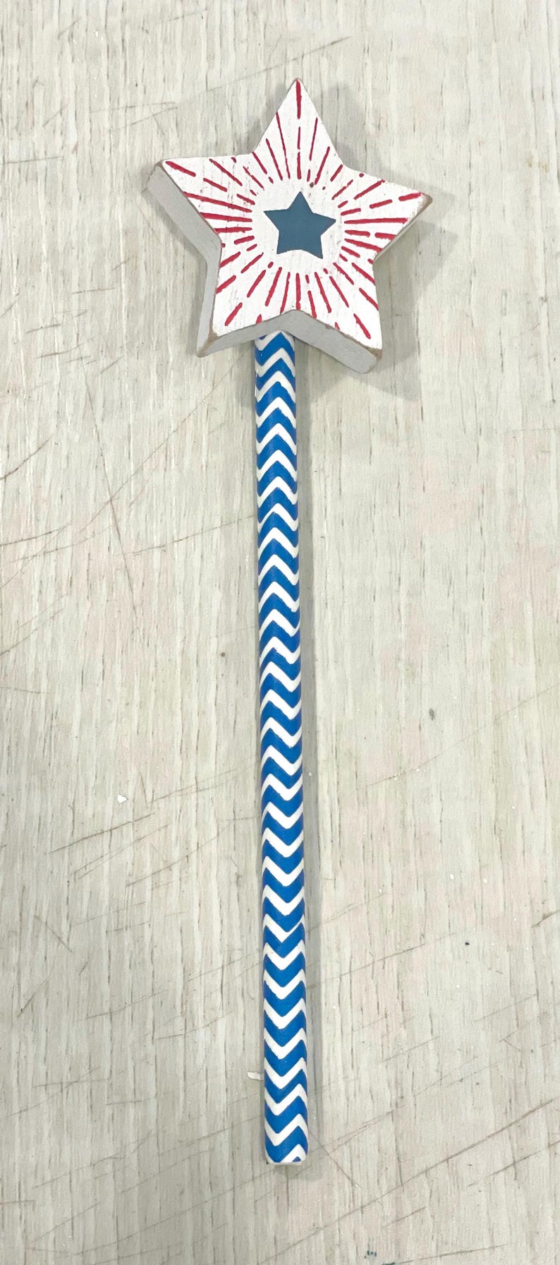 May include: A wooden star-shaped wand with a red and white starburst design and a blue star in the center. The wand has a blue and white chevron pattern.