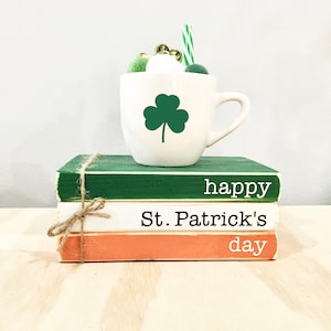 May include: Three stacked books with the words "happy", "St. Patrick's", and "day" written on them. A white mug with a green shamrock design sits on top of the books.