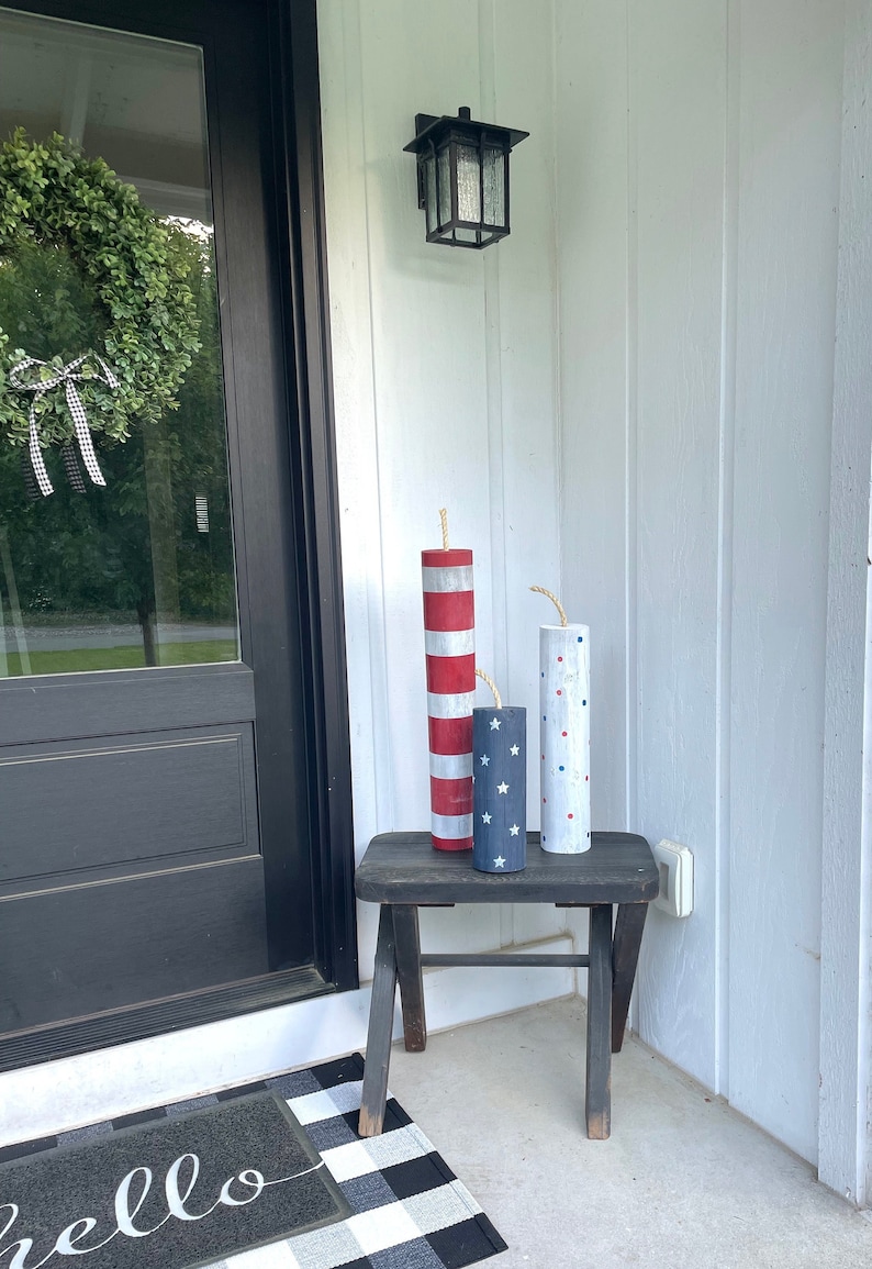 May include: Three wooden firework decorations painted in red, white, and blue with stars and polka dots. The decorations are sitting on a black wooden stool on a porch.