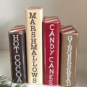 K&ouml;nnte beinhalten: Vier Holzbl&ouml;cke in verschiedenen Farben bemalt, die jeweils einem Buch &auml;hneln. Die Bl&ouml;cke sind mit "Hot Cocoa", "Marshmallows", "Candy Canes" und "Gingerbread" in wei&szlig;er Schrift beschriftet. Die Oberseiten der Bl&ouml;cke haben rot-wei&szlig; gestreifte Muster.