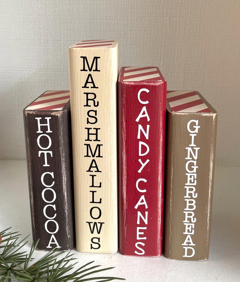 K&ouml;nnte beinhalten: Vier Holzbl&ouml;cke in verschiedenen Farben bemalt, die jeweils einem Buch &auml;hneln. Die Bl&ouml;cke sind mit "Hot Cocoa", "Marshmallows", "Candy Canes" und "Gingerbread" in wei&szlig;er Schrift beschriftet. Die Oberseiten der Bl&ouml;cke haben rot-wei&szlig; gestreifte Muster.